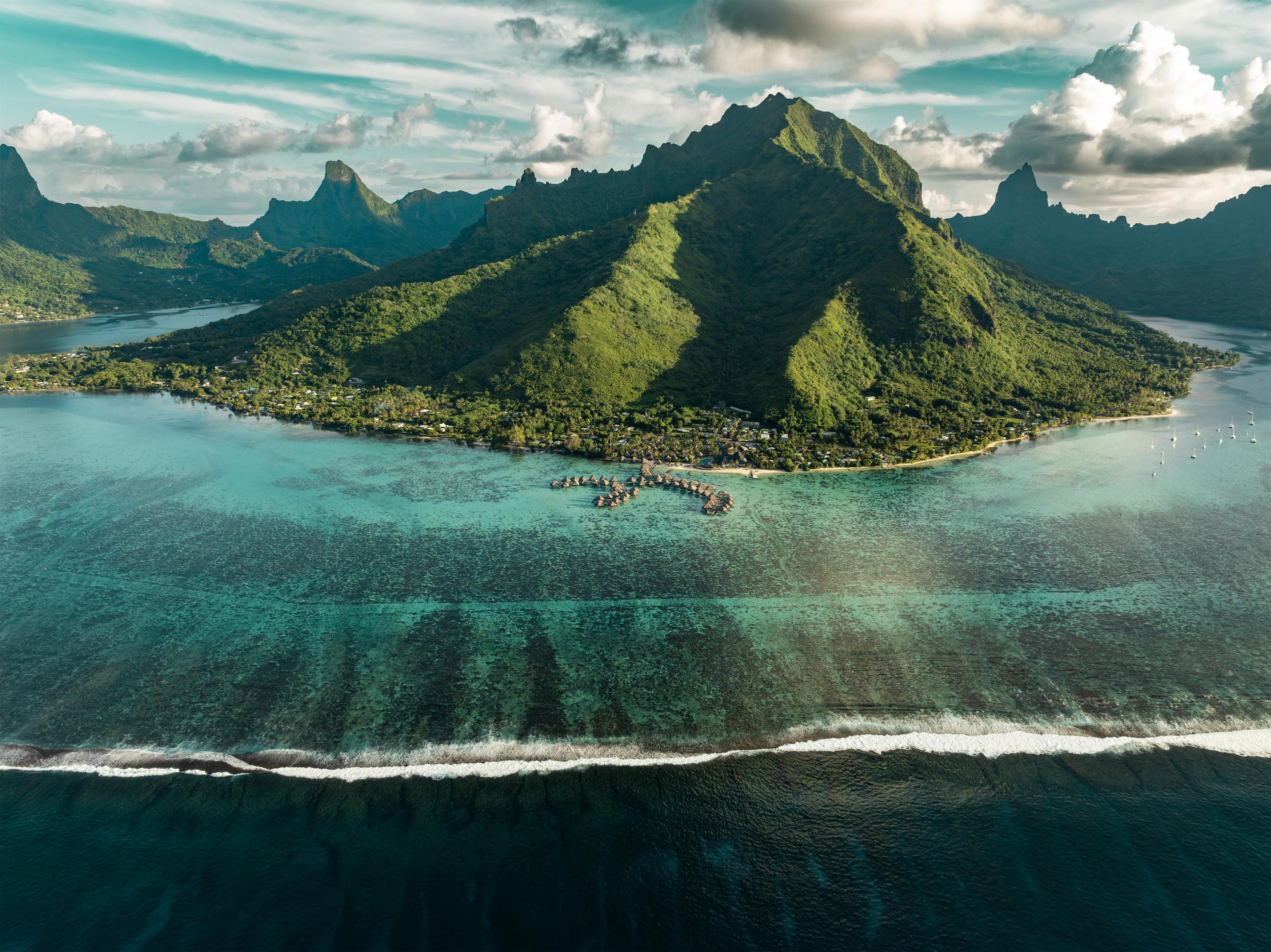 Vista aérea de una isla tropical con una montaña destacada, aguas turquesas cristalinas y un pequeño poblado con construcciones de techo de paja.