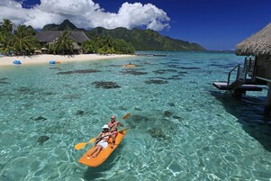 Un mar turquesa cristalino con una pareja haciendo kayak, una playa con palmeras y bungalows sobre el agua.