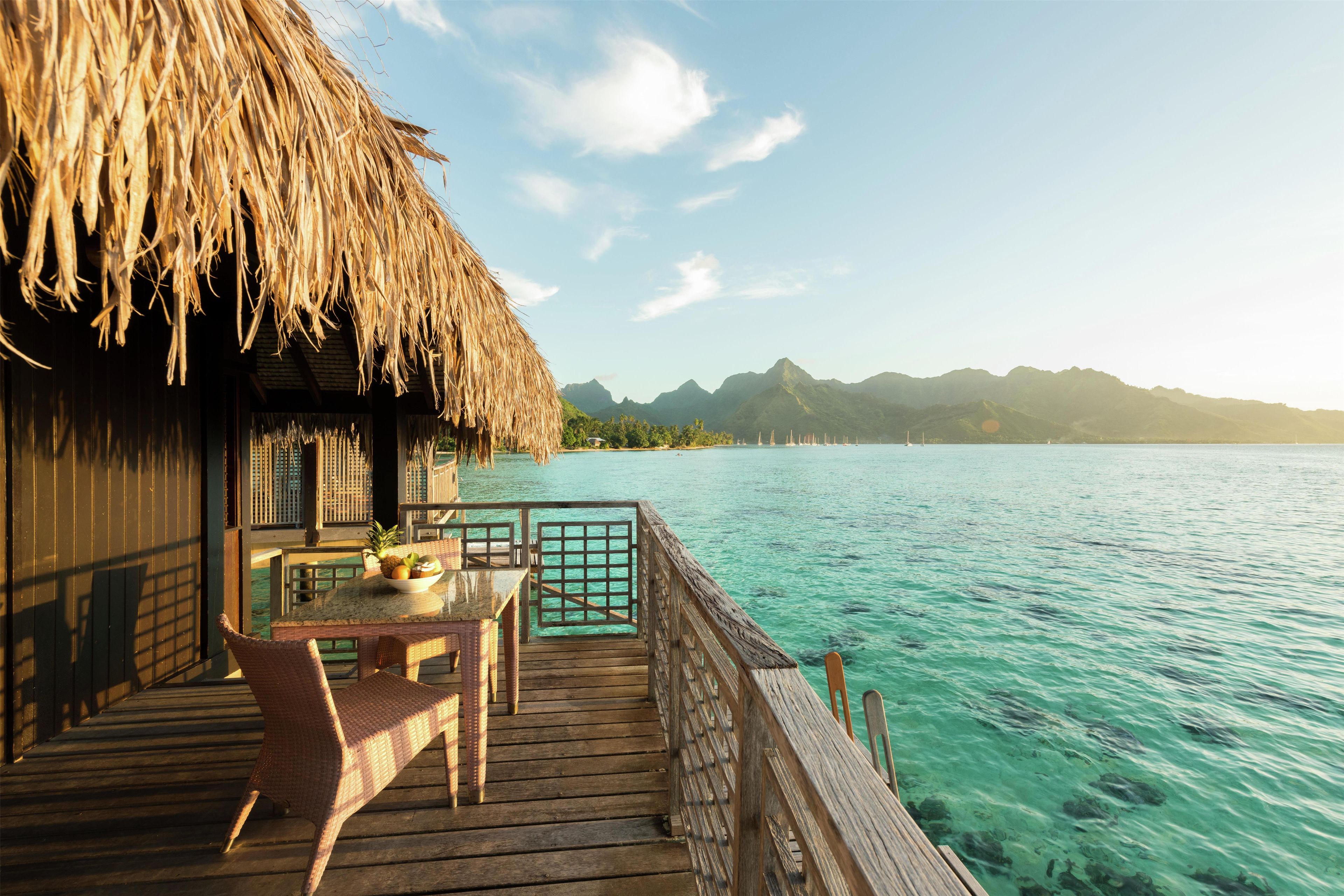 A wooden deck with a thatched roof, a table with chairs, and a view of clear turquoise water and distant mountains.