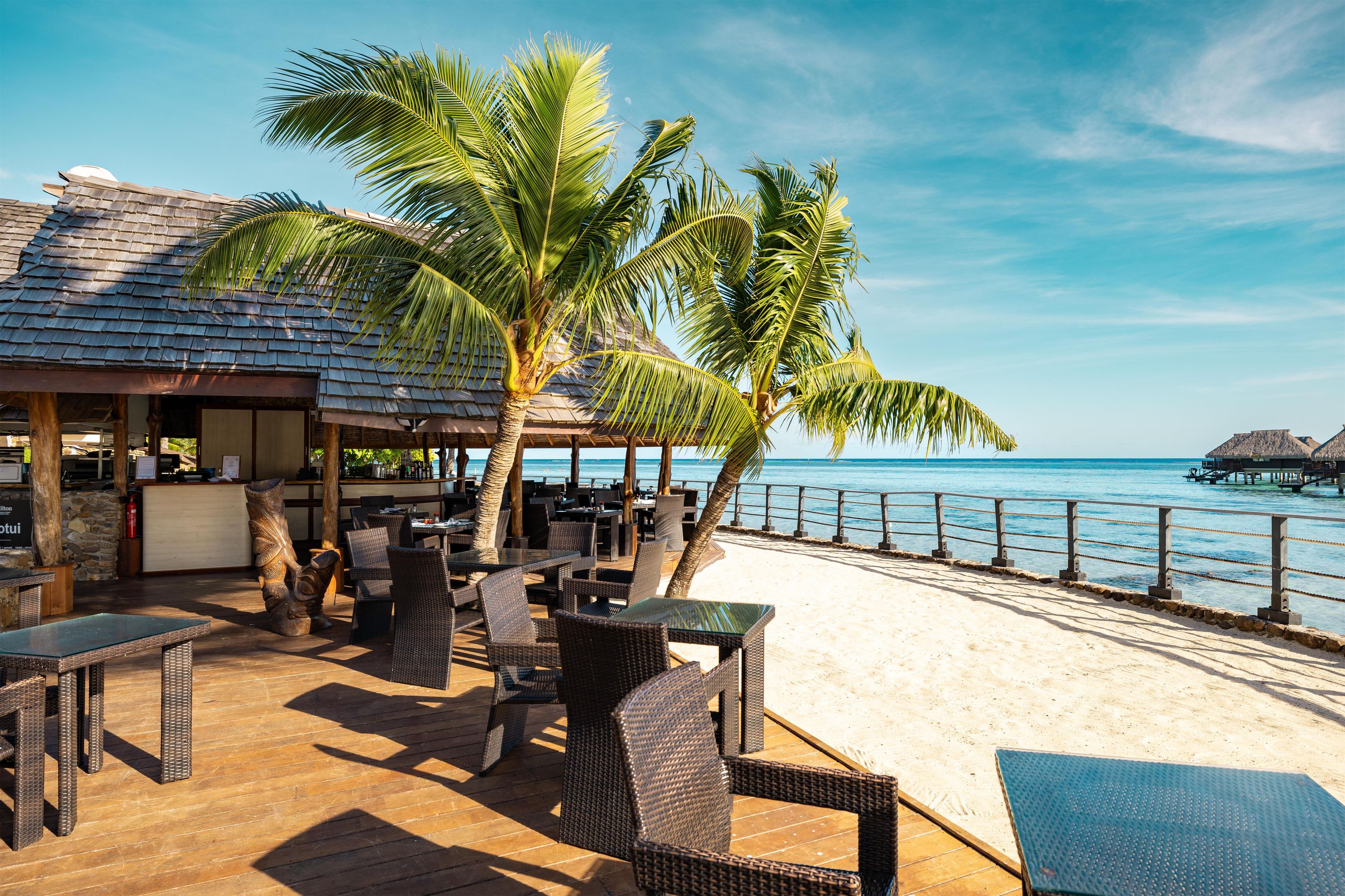 Een restaurant aan het strand met terras, palmbomen en uitzicht op de oceaan.
