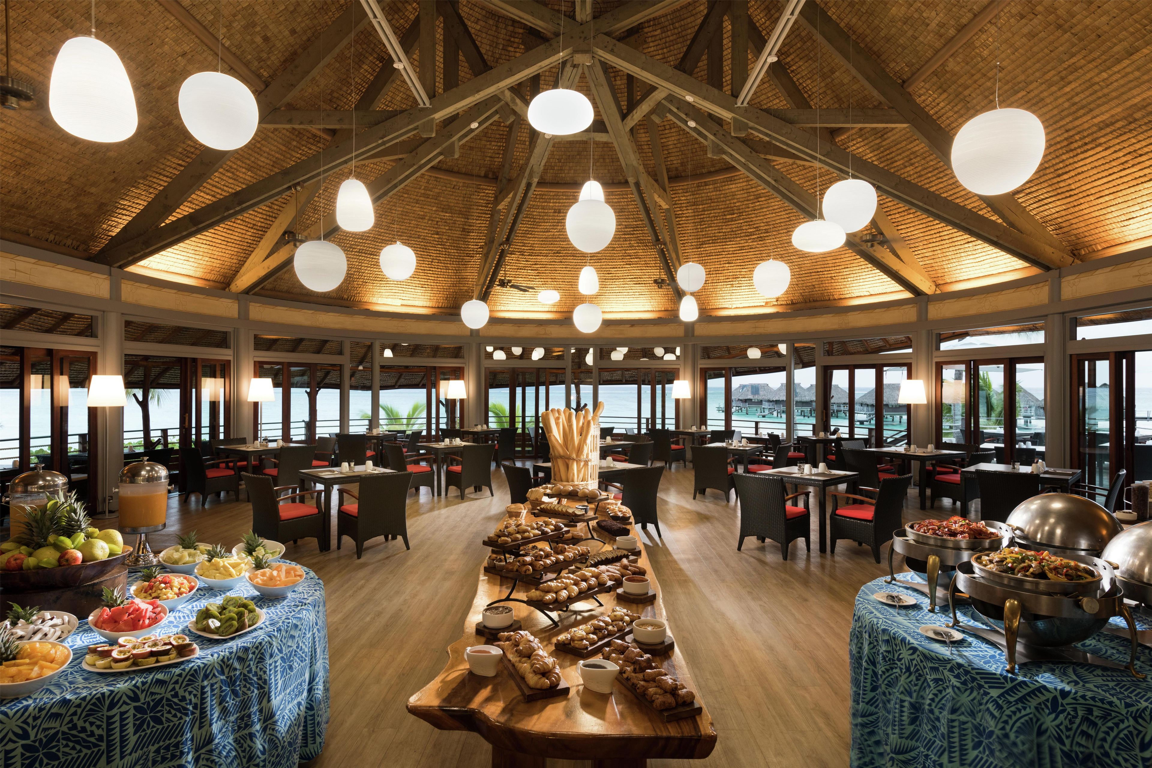 A spacious dining area with a long table set for breakfast, large windows, and a high ceiling with exposed beams.