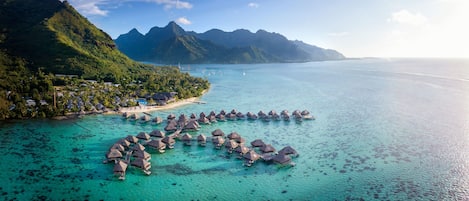 Vista aérea de un resort tropical con bungalows sobre el agua, una playa y montañas exuberantes al fondo.