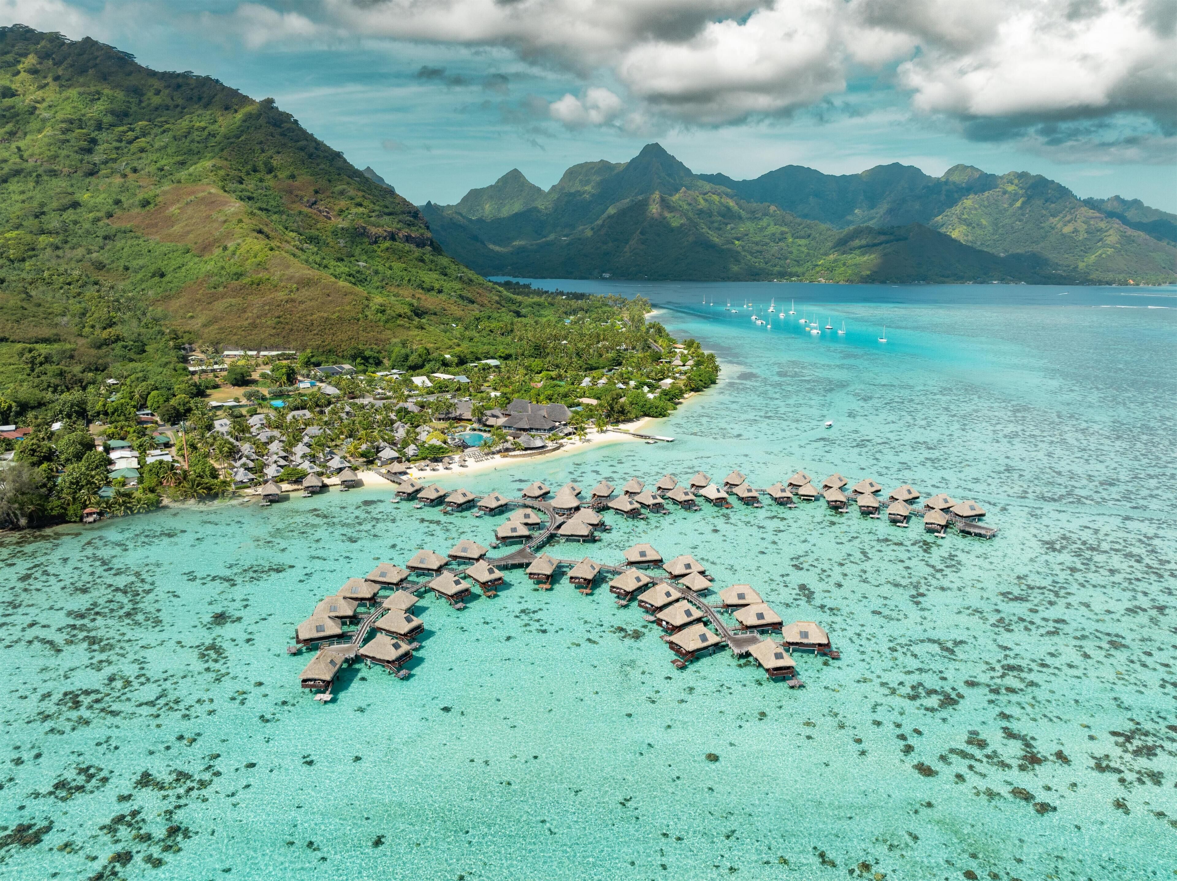 Une vue aérienne de bungalows sur pilotis disposés en arc de cercle au-dessus d’un récif turquoise, entourés d’une végétation luxuriante et de montagnes.