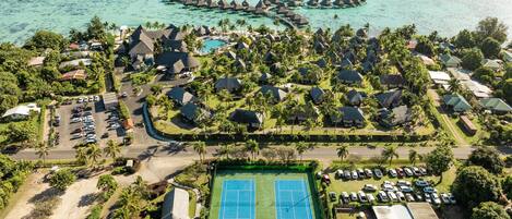 An aerial view of a resort with overwater bungalows, a tennis court, and a parking area.