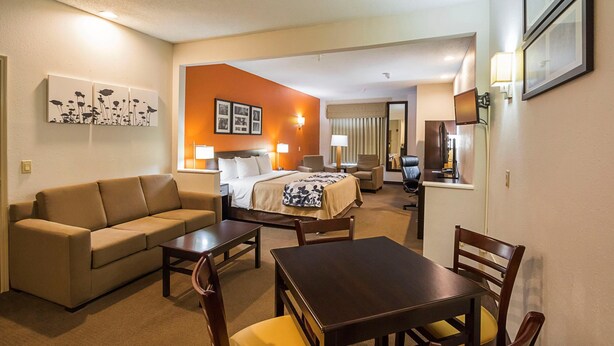 A hotel room with a sofa, a bed, a table, and chairs. There are wall-mounted lamps and framed pictures. - Sleep Inn And Suites Danville (Danville)