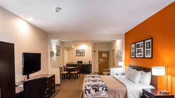 A hotel room with a bed, desk, chair, television, and a kitchenette in the background.
