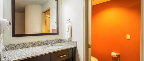 A bathroom with a granite countertop sink, dark cabinetry, a large mirror, and a toilet against an orange wall.