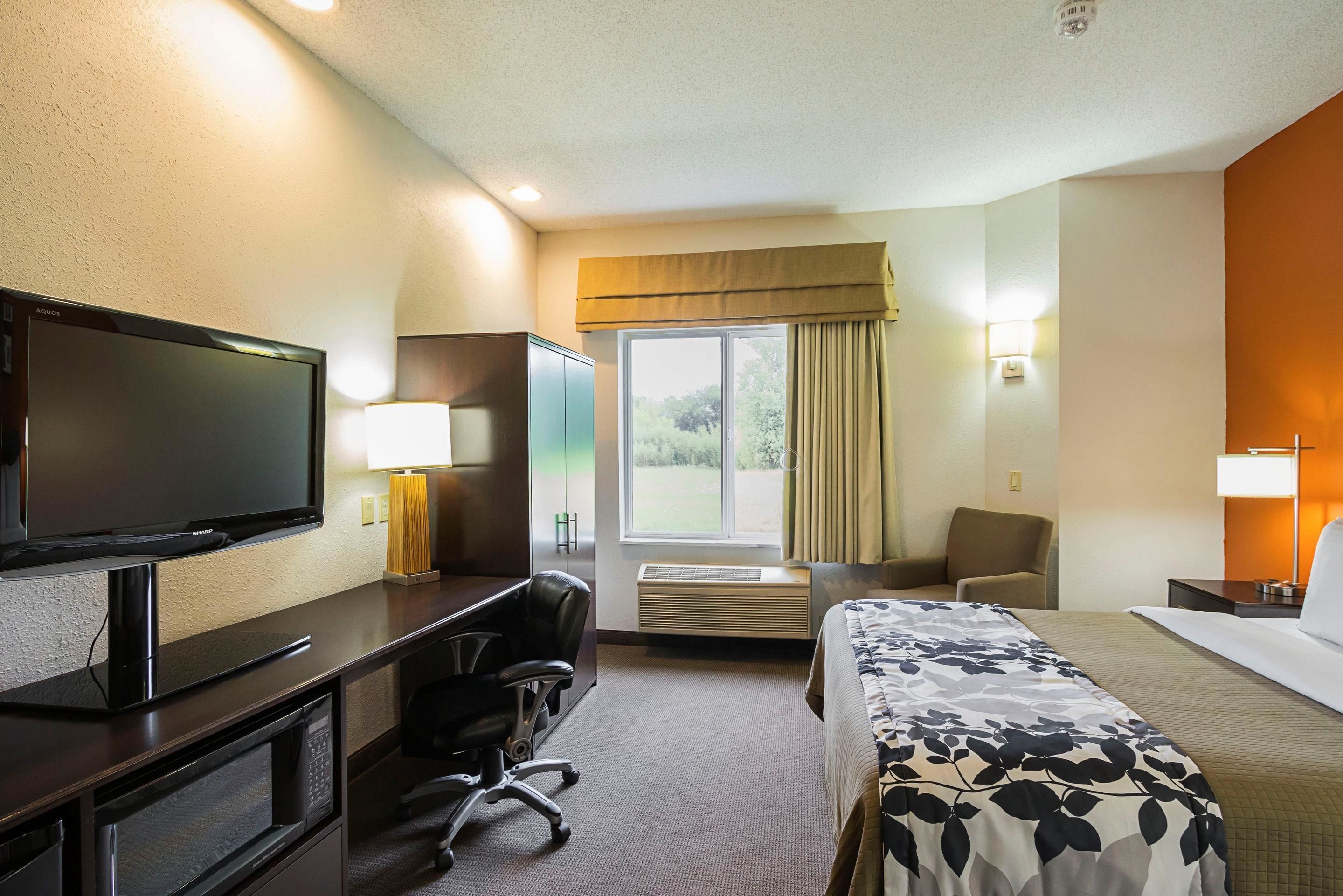 Habitación de hotel con cama, escritorio con televisor, silla, ventana con cortinas y lámpara fijada a la pared.