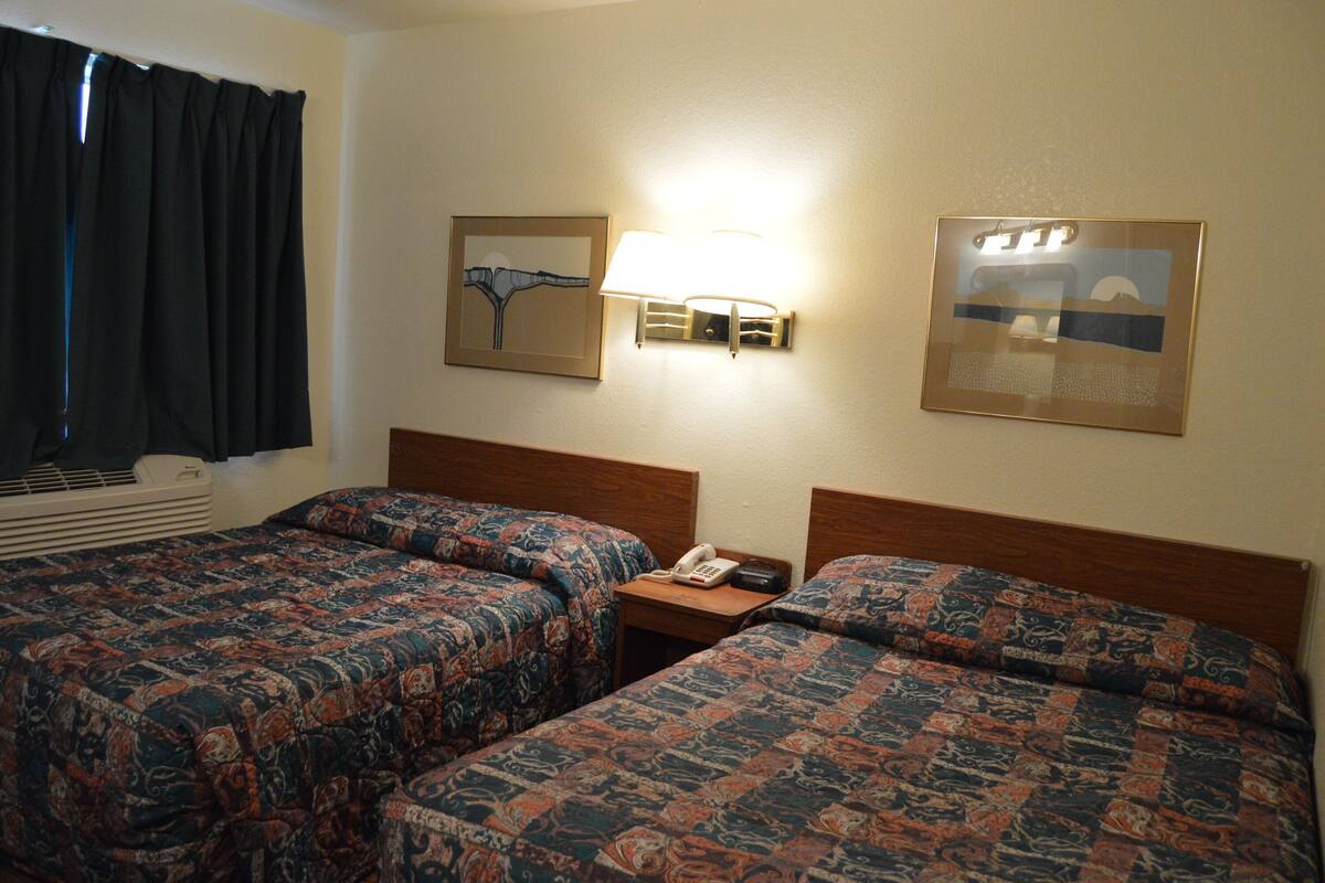 A hotel room with two beds, a nightstand with a phone, a wall-mounted lamp, and two framed pictures.