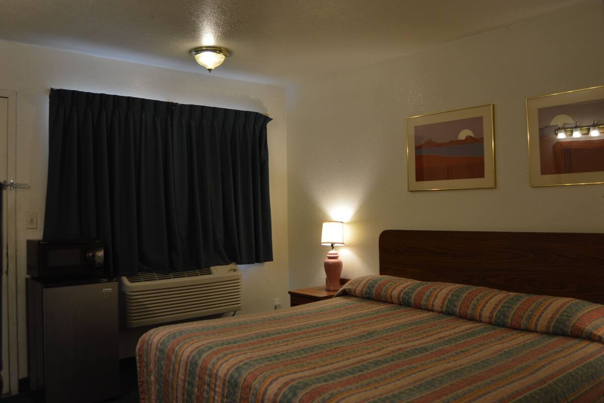 A hotel room with a bed, a desk with a computer, a lamp, and two framed pictures on the wall.