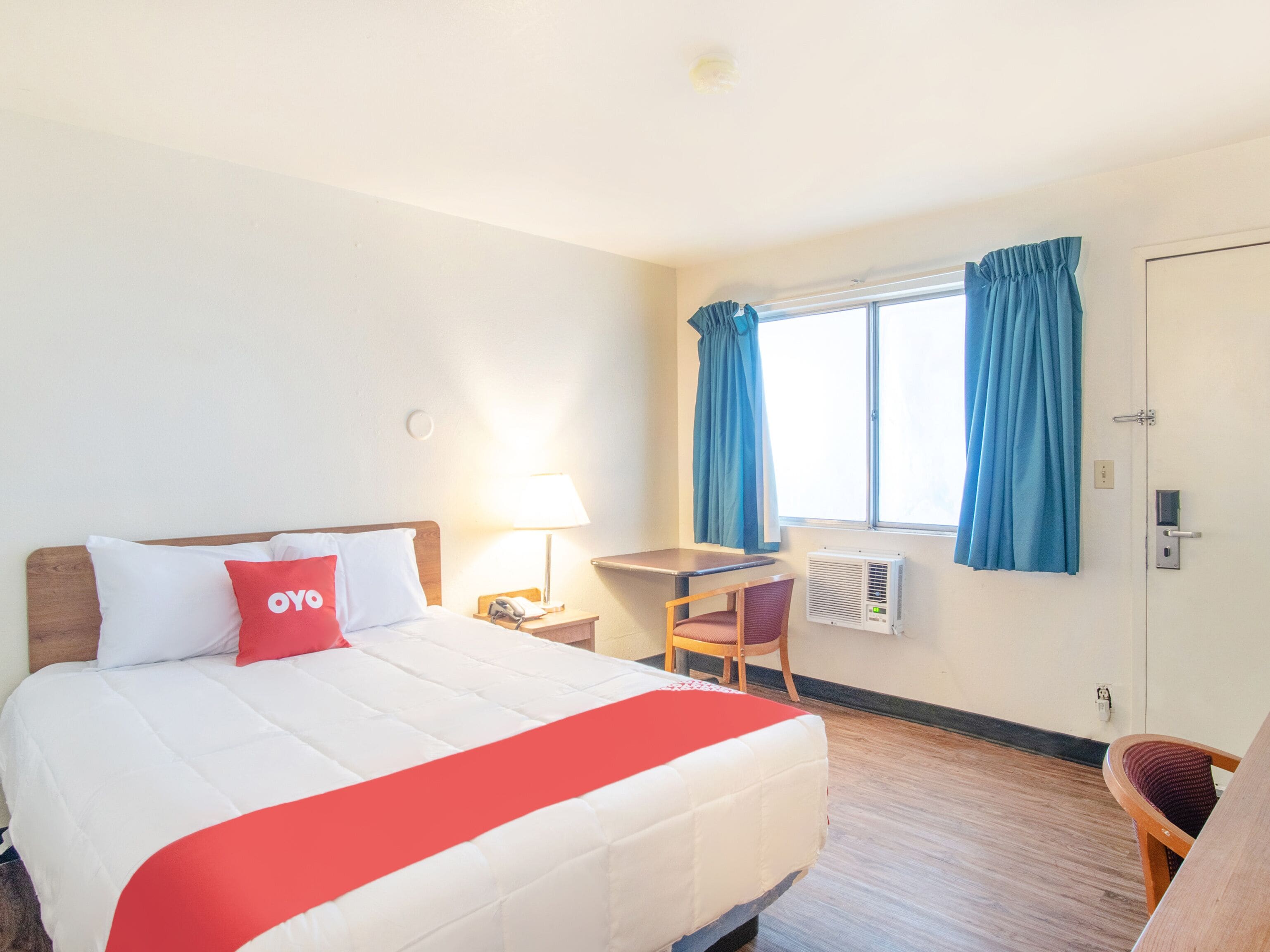 Ein Hotelzimmer mit einem Bett, einem Schreibtisch, einem Stuhl, einem Fenster mit Vorhängen und einer Klimaanlage.