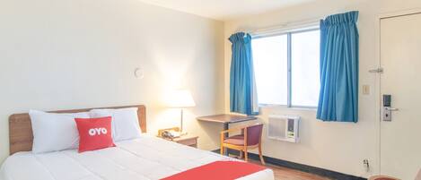 Ein Hotelzimmer mit einem Bett, einem Schreibtisch, einem Stuhl, einem Fenster mit Vorhängen und einer Klimaanlage.