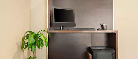 A desk with a computer monitor, keyboard, and printer, a potted plant, and a wall-mounted shelf.