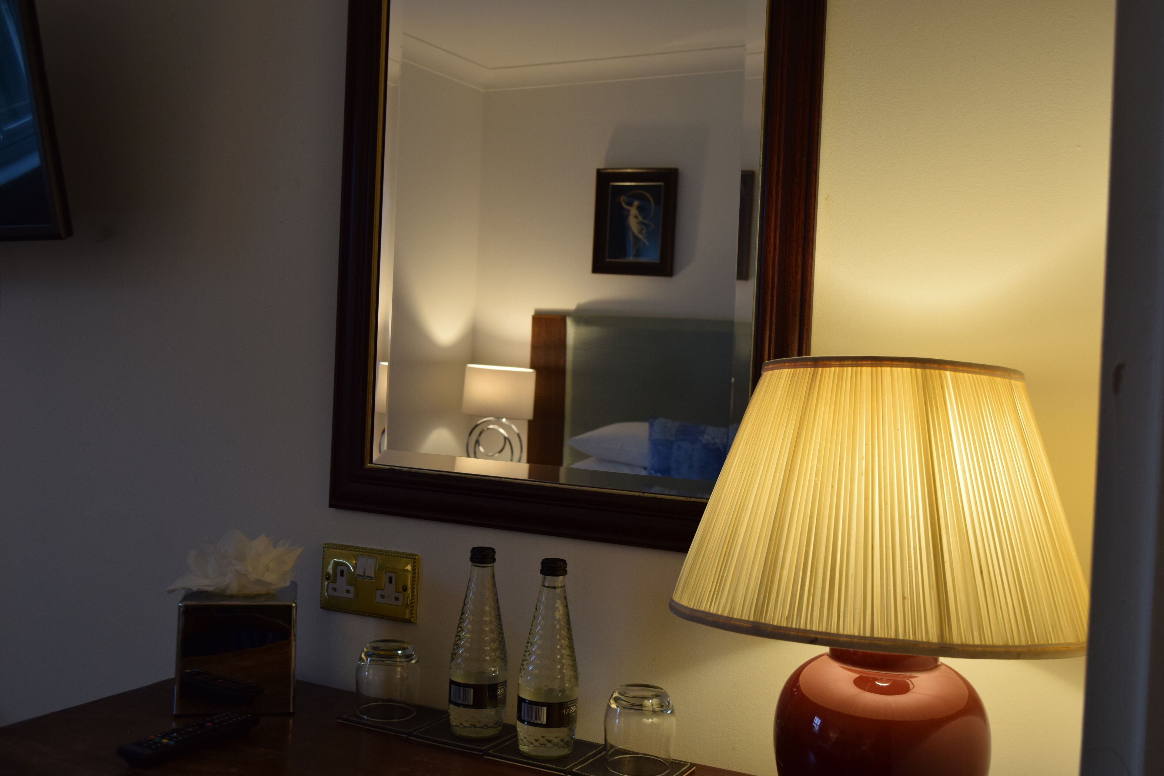 Habitación de hotel con una lámpara de noche, un espejo que refleja una cama y un cuadro, y una mesita con una caja de pañuelos, dos botellas y un control remoto.