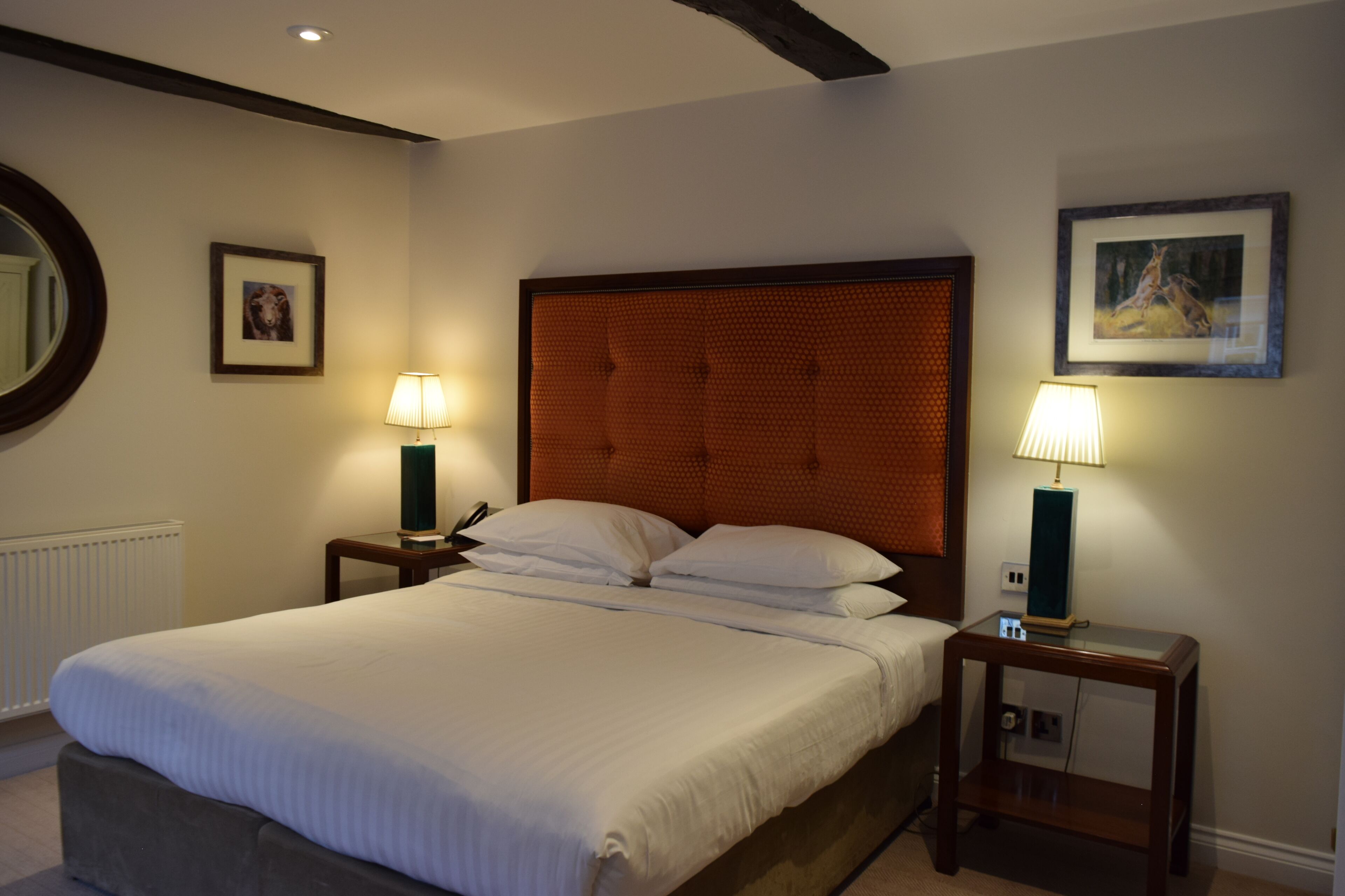A neatly made bed with white linens, two bedside tables with lamps, a framed picture on the wall, and a round mirror on the left.
