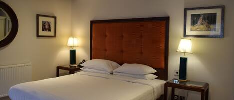 A neatly made bed with white linens, two bedside tables with lamps, a framed picture on the wall, and a round mirror on the left.