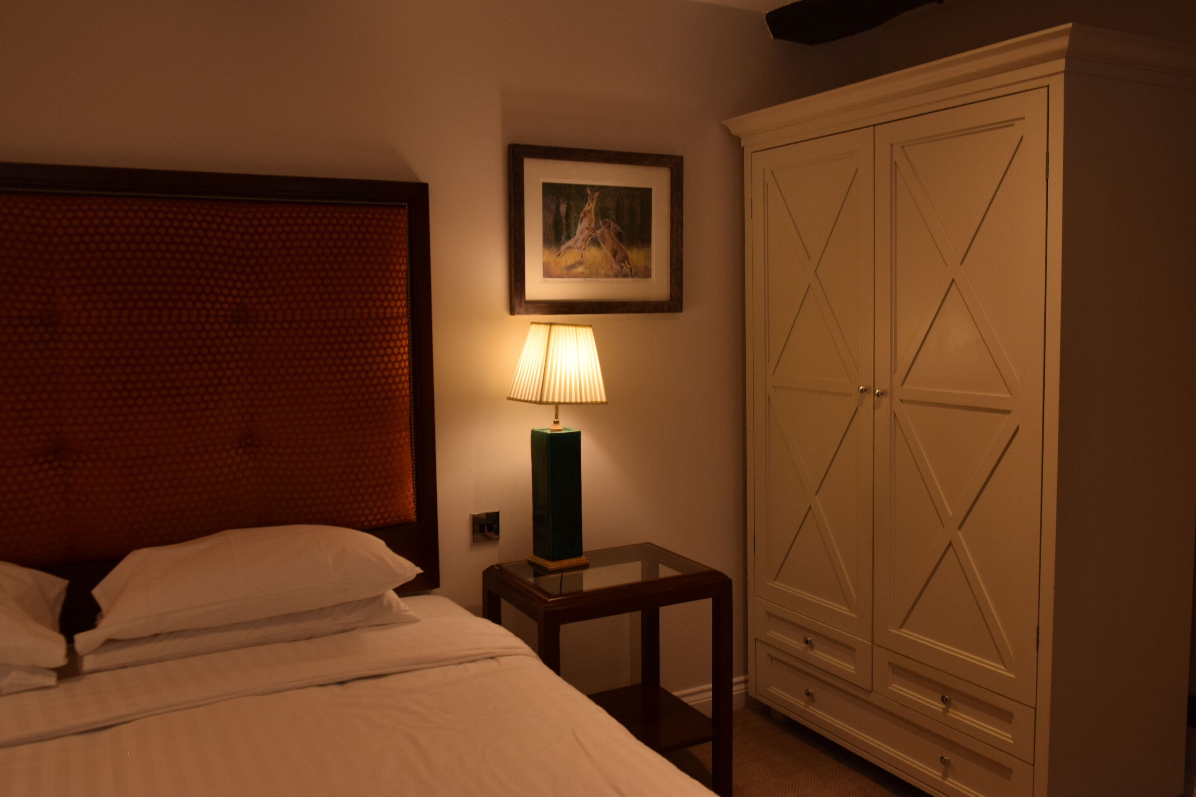 A bedroom with a bed, a bedside table with a lamp, a wardrobe, and a framed picture on the wall.