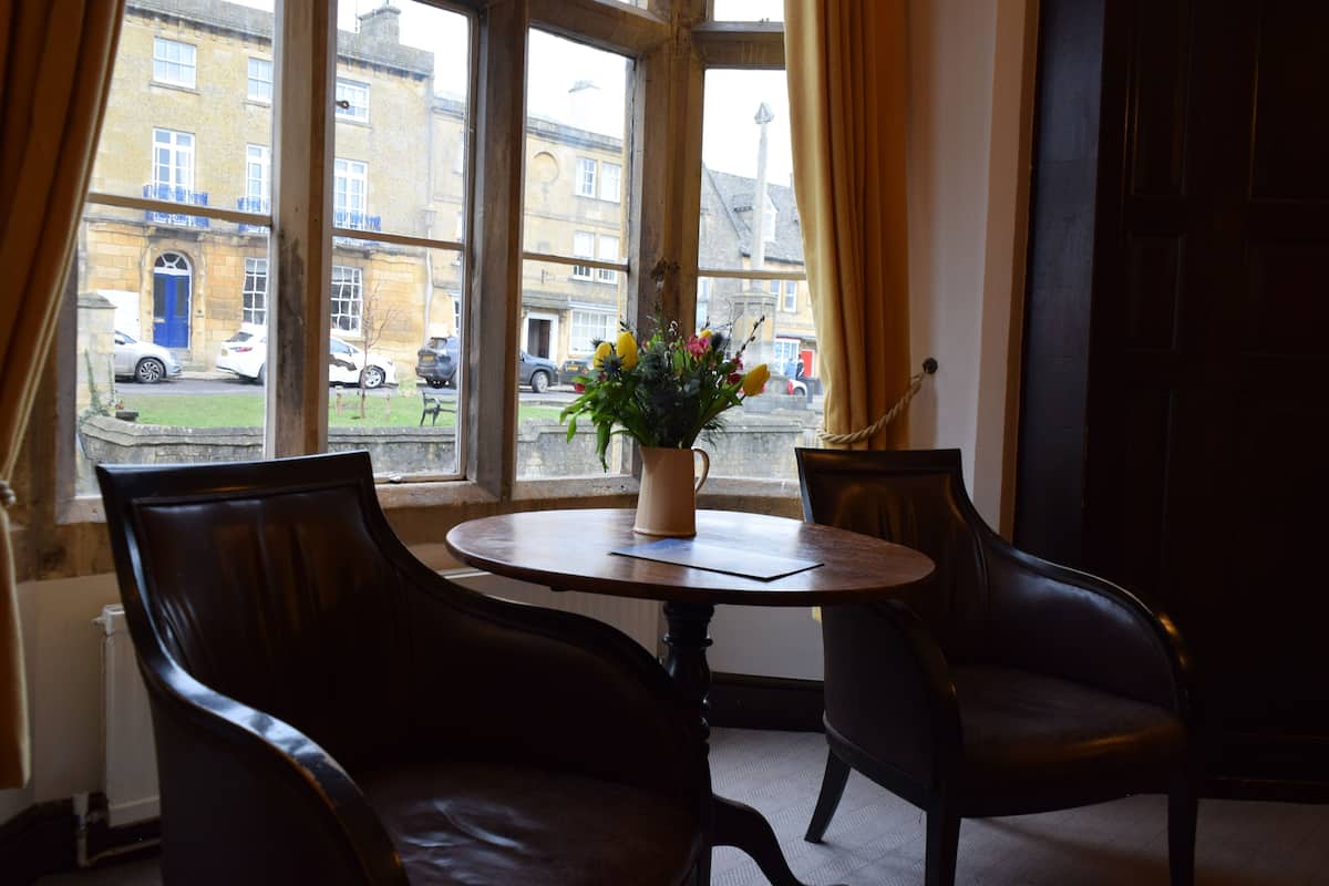 A room with a round table, two leather chairs, a vase with flowers, and a view of a street with parked cars and buildings.