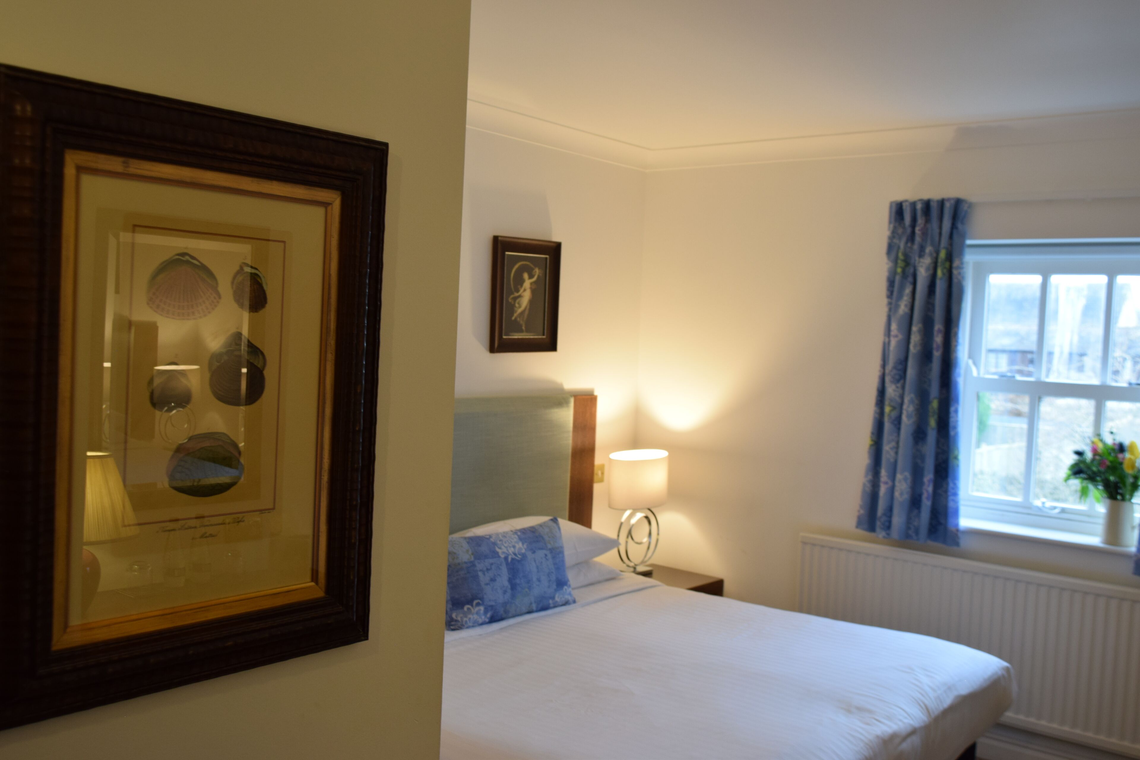 A hotel room with a bed, a framed picture on the wall, a bedside lamp, and a window with curtains.