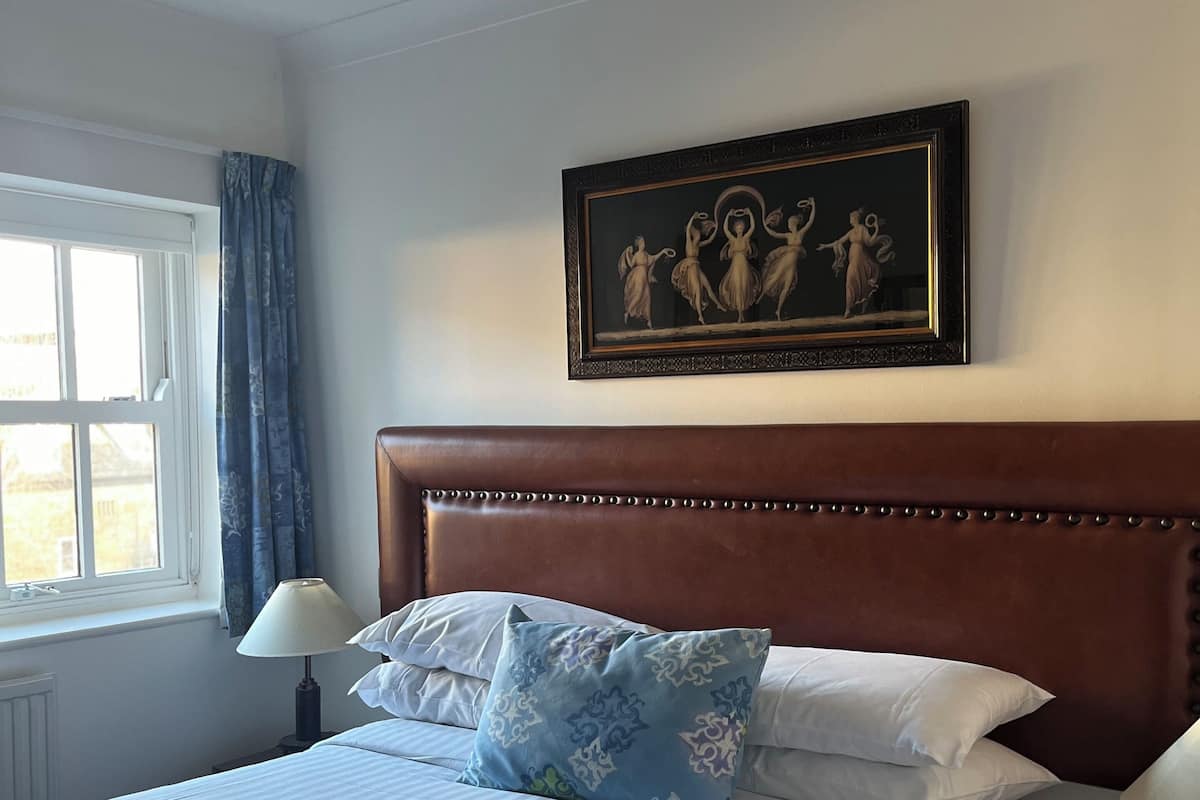 A neatly made bed with white linens and a blue patterned pillow, a wooden headboard, a framed artwork above the bed, and a window with curtains.