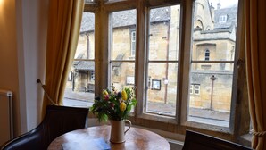 A round wooden table with a vase of flowers, two chairs, and a view of historic buildings through large windows with curtains. - Noel Arms Hotel (Chipping Campden)