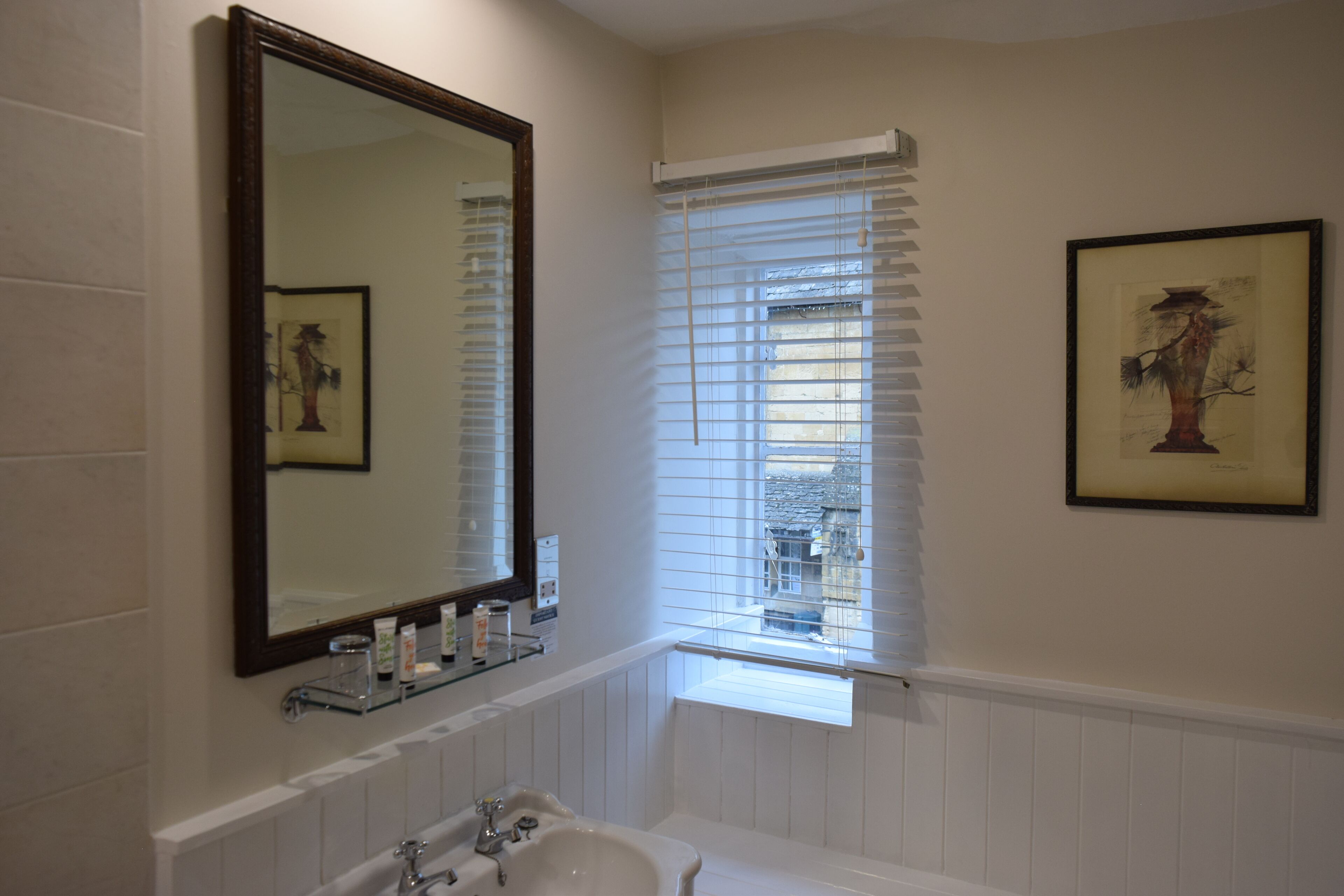 A bathroom with a large mirror, a window with blinds, a framed picture on the wall, and a sink with a glass shelf above it.