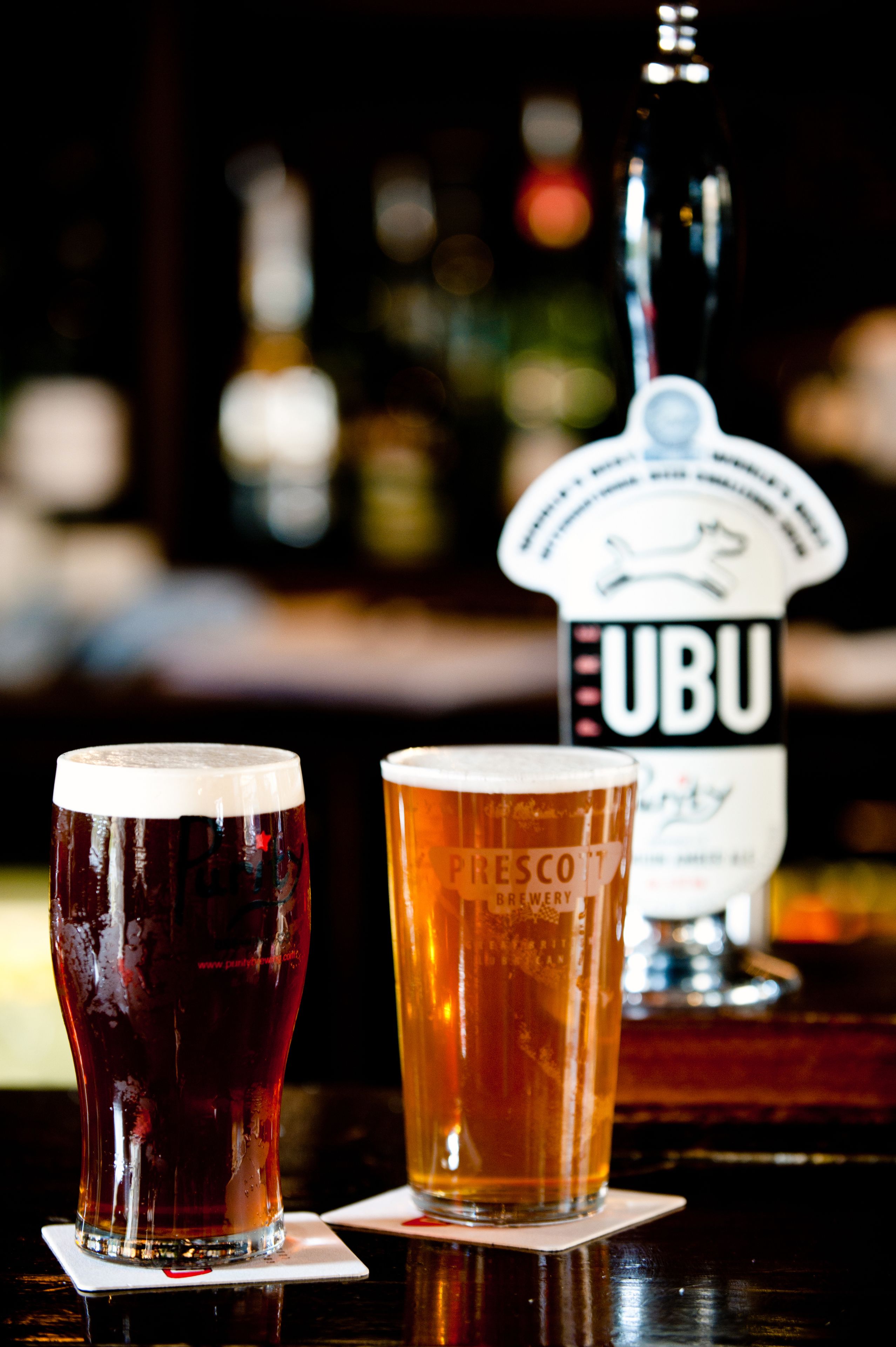 Two glasses of beer on a bar counter with a beer tap labeled "UBU" in the background.
