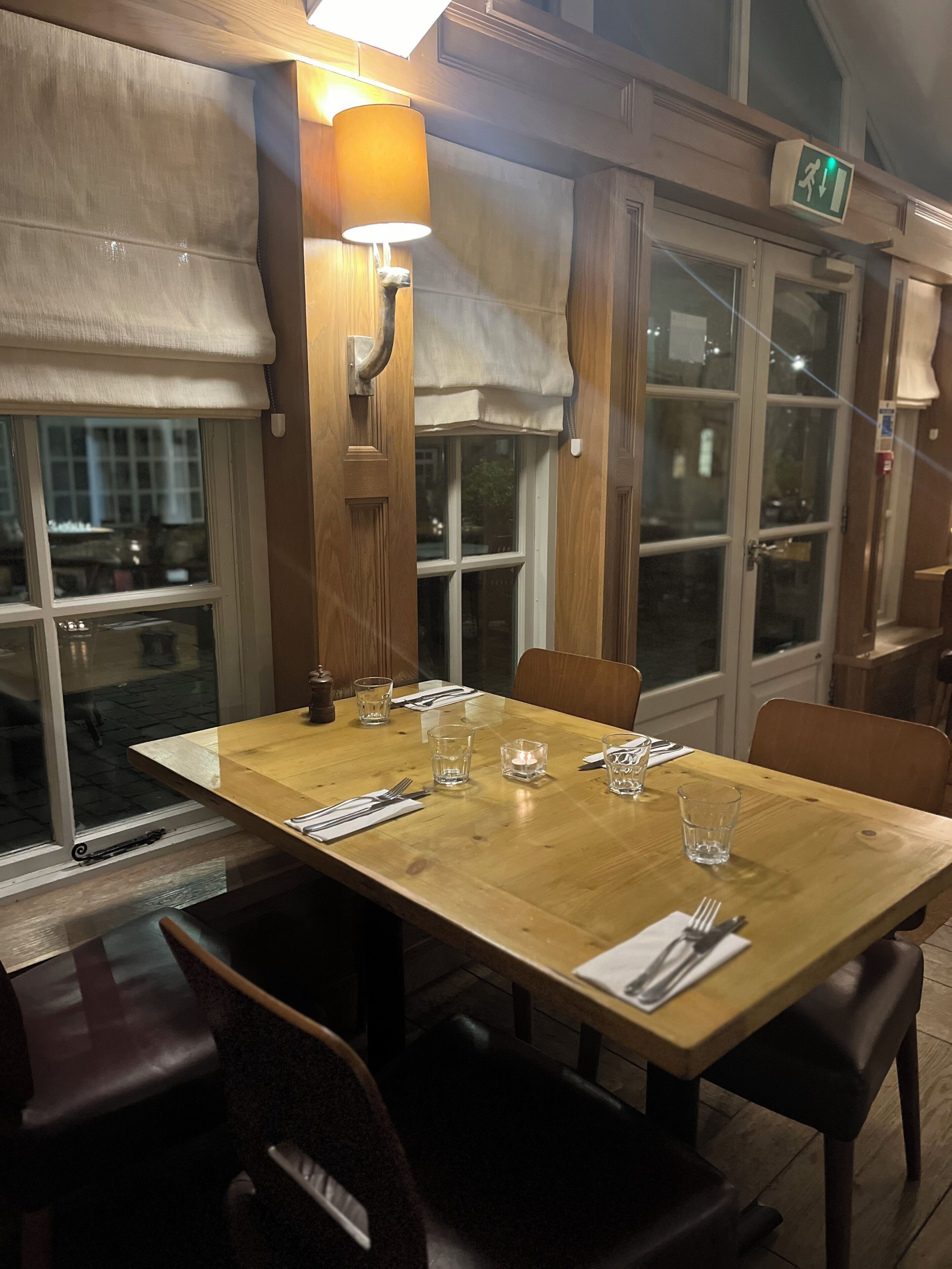 Uma sala de jantar com uma mesa de madeira posta para quatro pessoas, incluindo copos e talheres. Há duas cadeiras, um abajur e uma janela com vista para a rua.