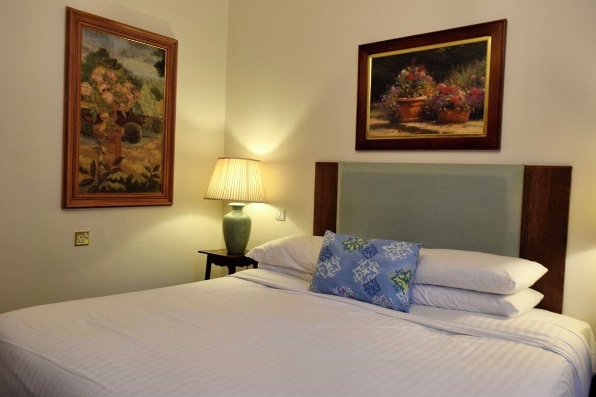 Una cama bien tendida con sábanas blancas y una almohada de estampado azul, una lámpara de cerámica verde en una mesita de noche, y dos cuadros con motivos florales en la pared.