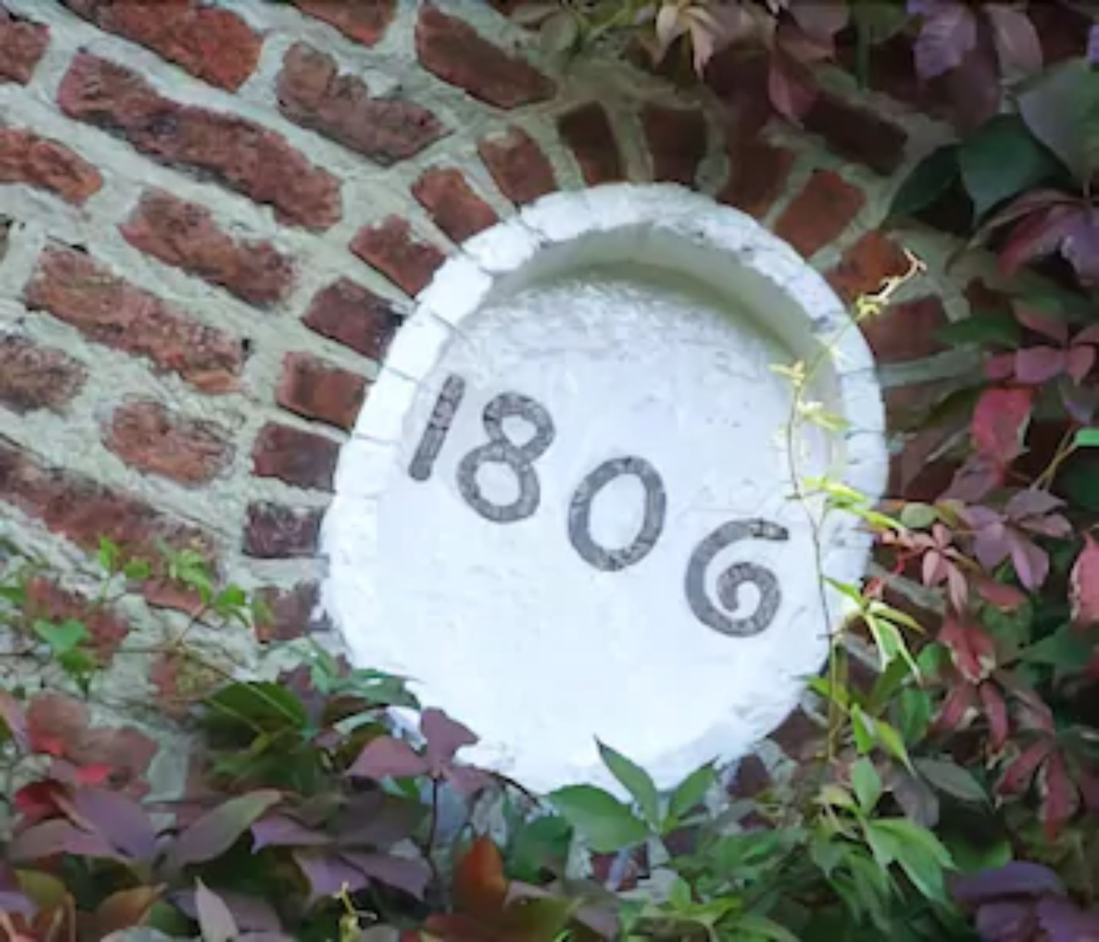 A white stone plaque with the number "1806" on a brick wall, surrounded by foliage.