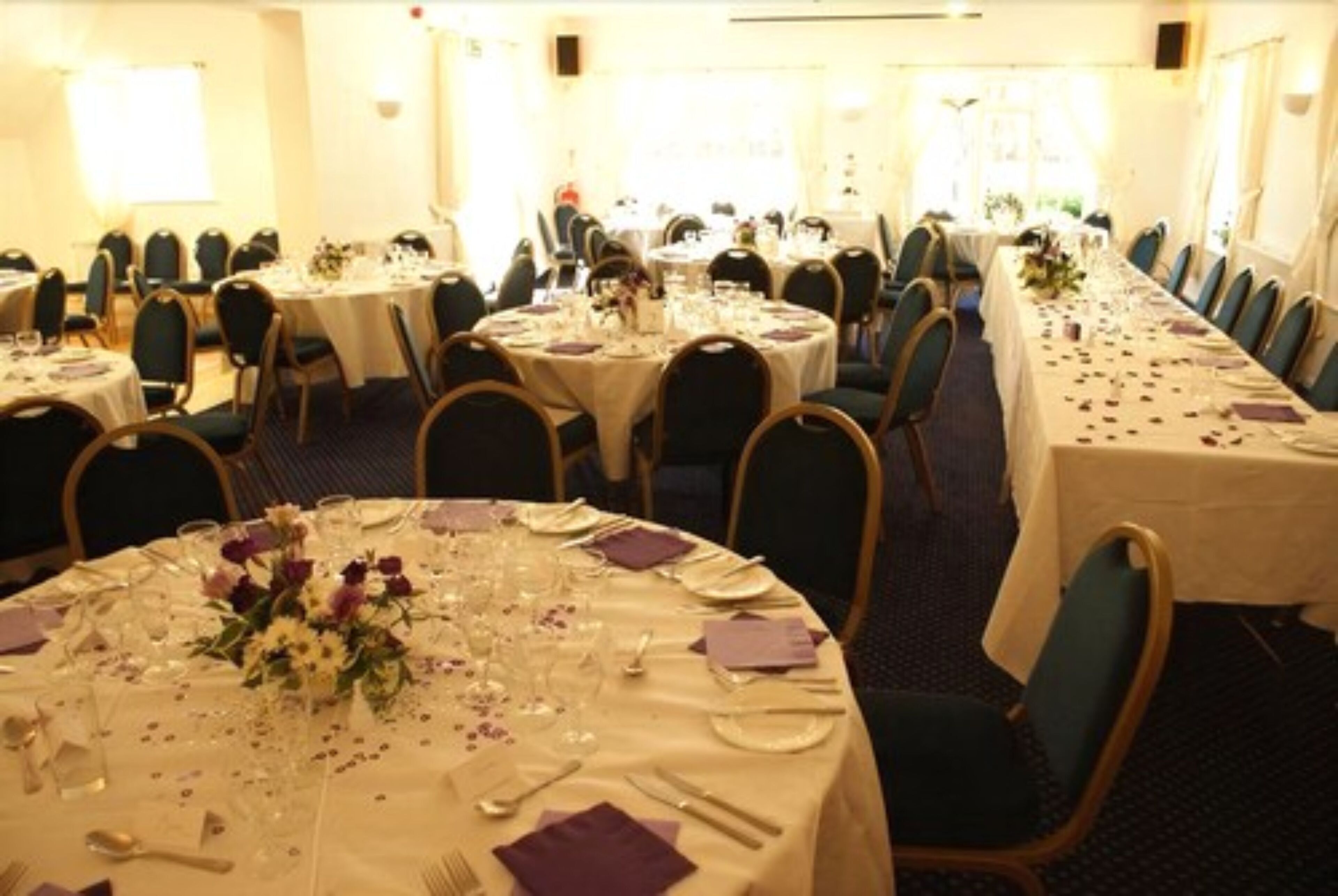A banquet hall with multiple round tables set for dining, each with white tablecloths, chairs, and floral centerpieces.