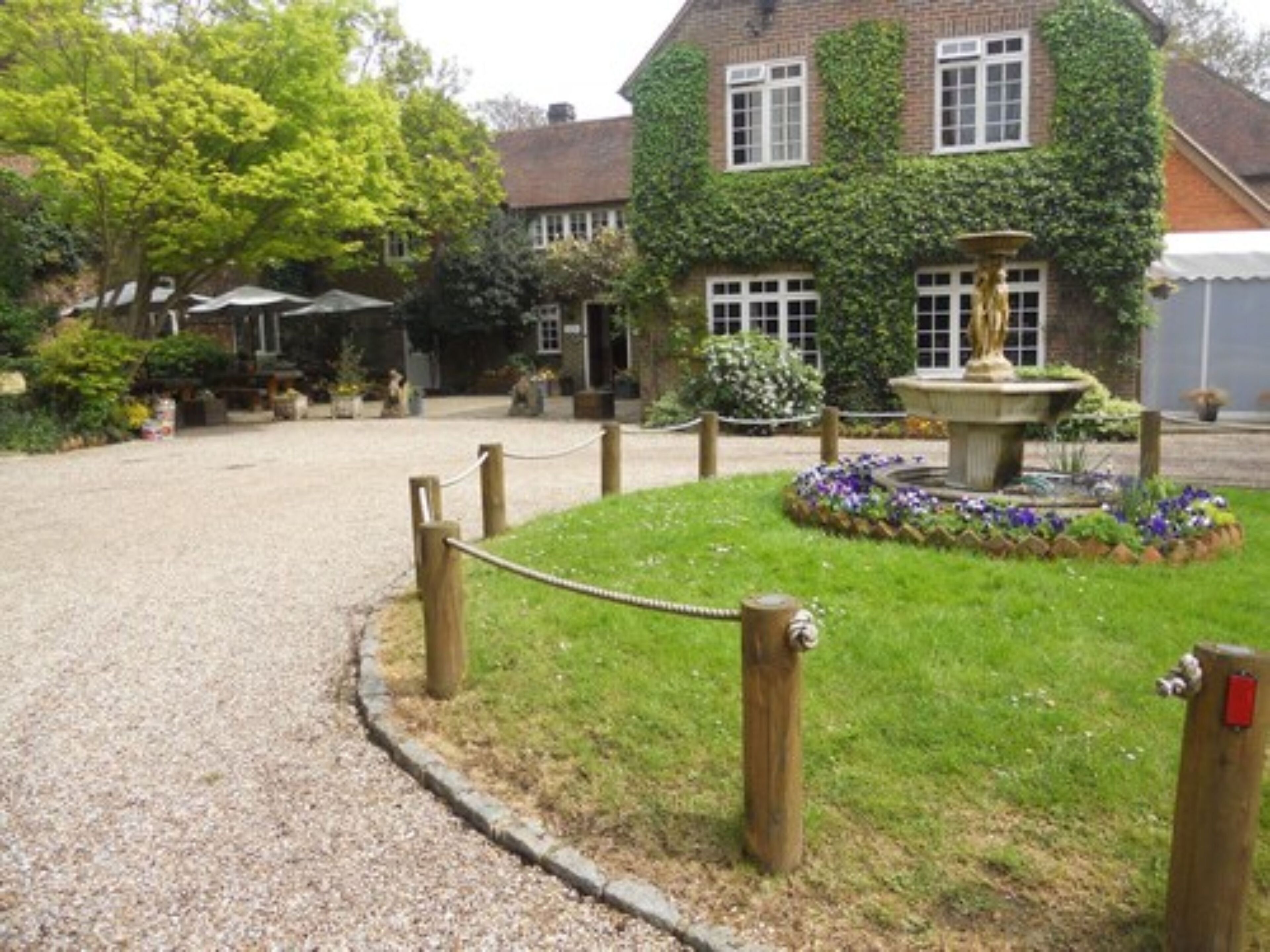A building with ivy-covered walls, a gravel driveway, and a fountain surrounded by flowers.