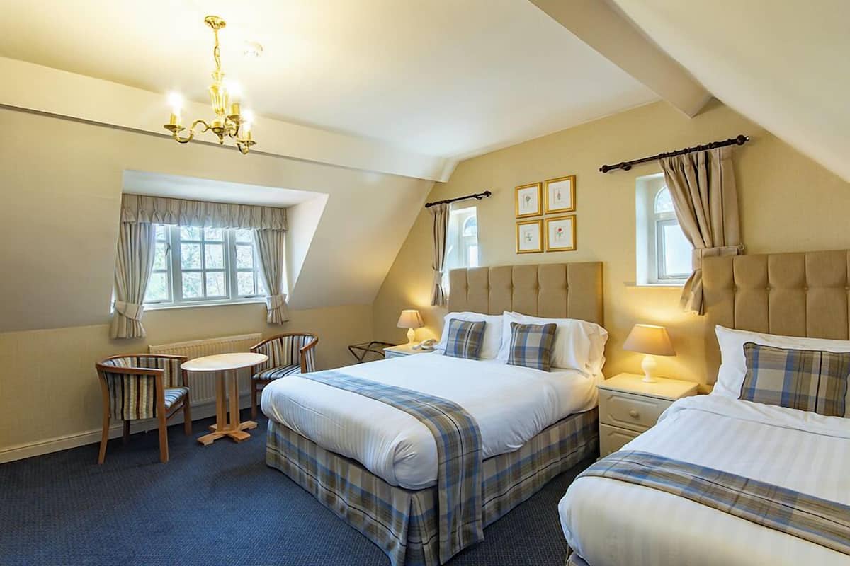 A hotel room with a large bed, a smaller bed, a desk with a chair, a chandelier, and a window with curtains.