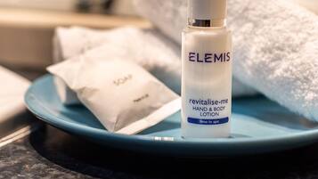Une bouteille de lotion pour les mains et le corps « Revitalise Me » de la marque Elemis, posée sur une assiette bleue, avec une serviette blanche et un petit sachet de savon en arrière-plan.