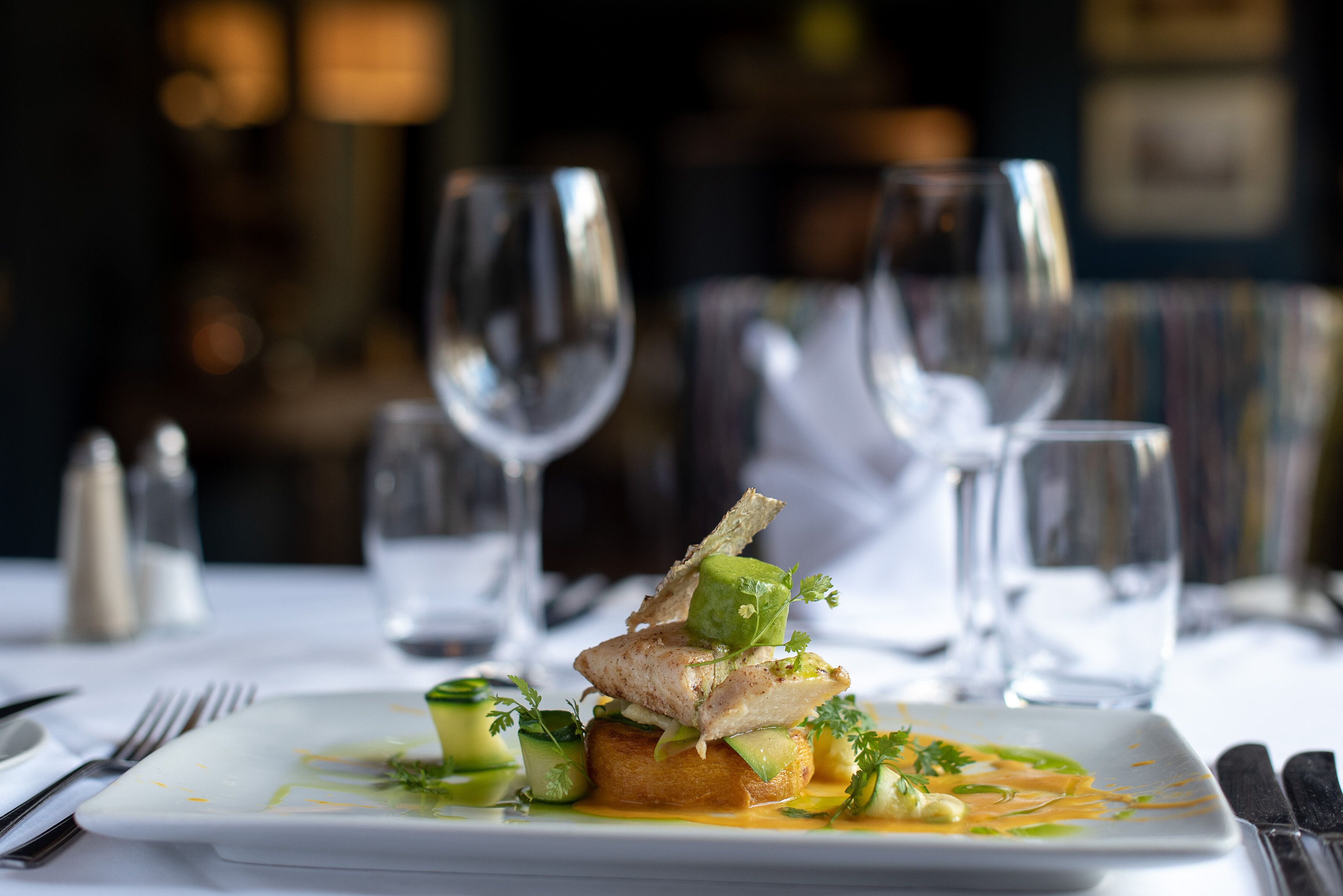 Un plato cuadrado blanco con un plato de pescado, adornado con hierbas y verduras, sobre un mantel blanco.