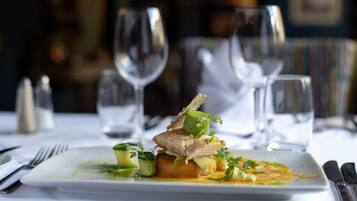 Un plato cuadrado blanco con un plato de pescado, adornado con hierbas y verduras, sobre un mantel blanco.