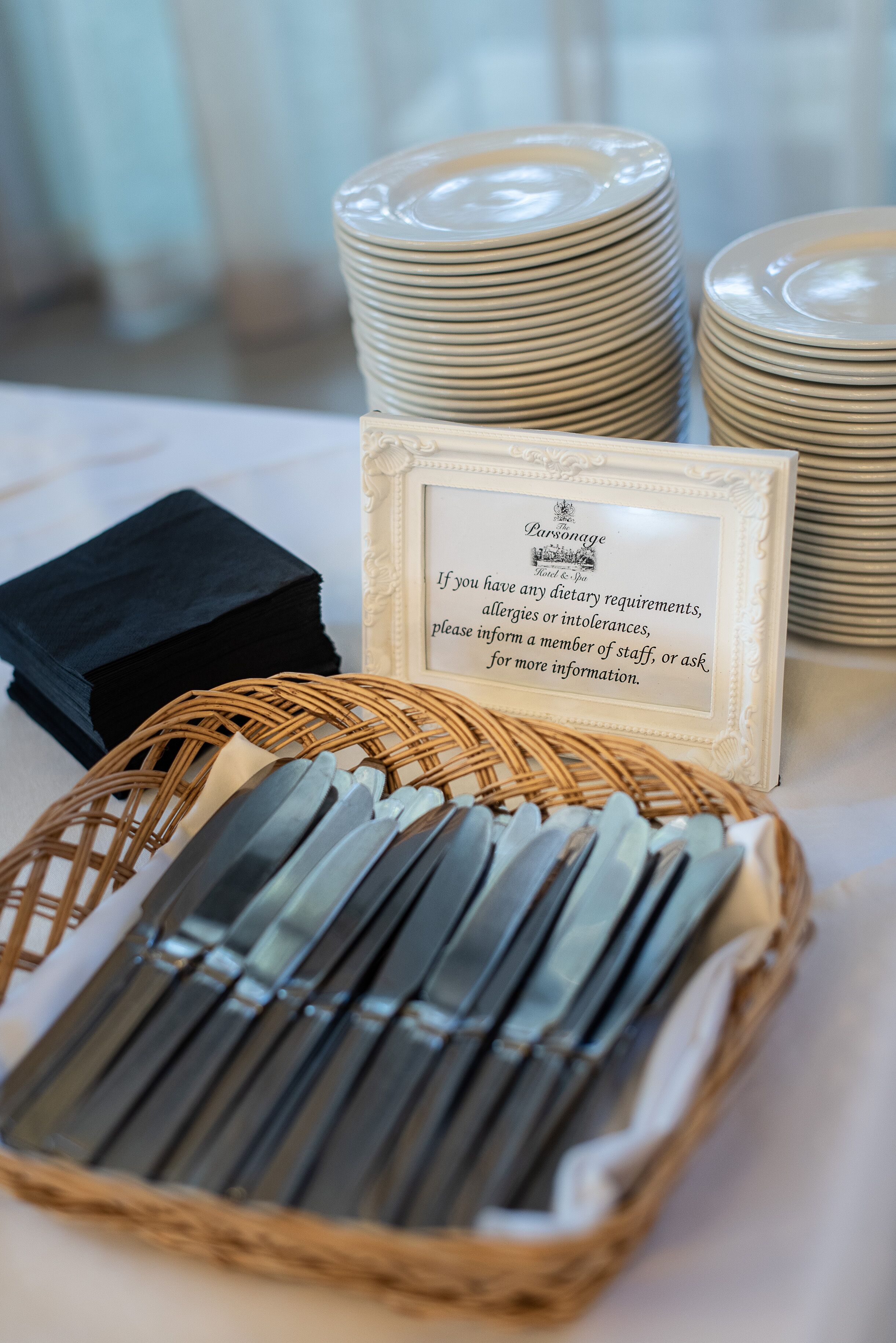A stack of white plates, a wicker basket with silverware, and a sign with dietary requirements information.