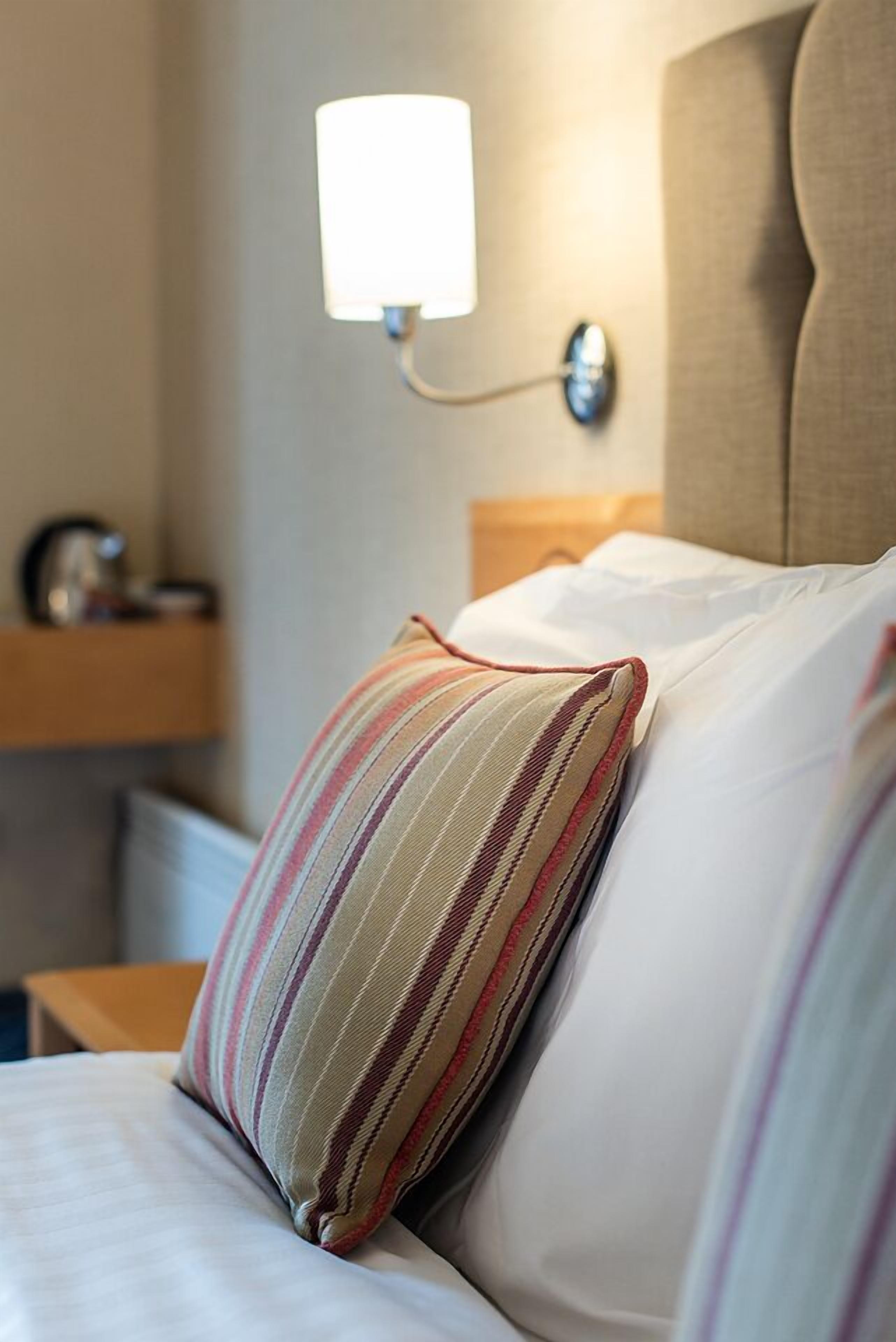 Een hotelkamer met een bed, een gestreept kussen, een wandlamp en een nachtkastje van hout met een klok.