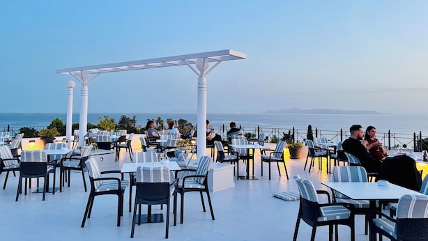 Un área de comedor al aire libre con mesas y sillas, una pérgola y vista al mar.