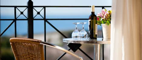 A wicker chair and a table with a bottle and glasses on a balcony with a metal railing and a view of the ocean.