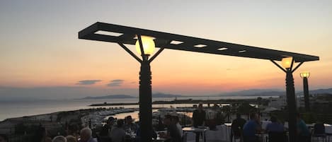 An outdoor dining area with tables and chairs, a pergola structure, and a view of the sea and distant mountains during sunset.