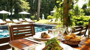 Mesa de comedor con platos de comida, vasos y una cesta de pan, con vistas a una piscina con sillas de descanso y sombrillas.