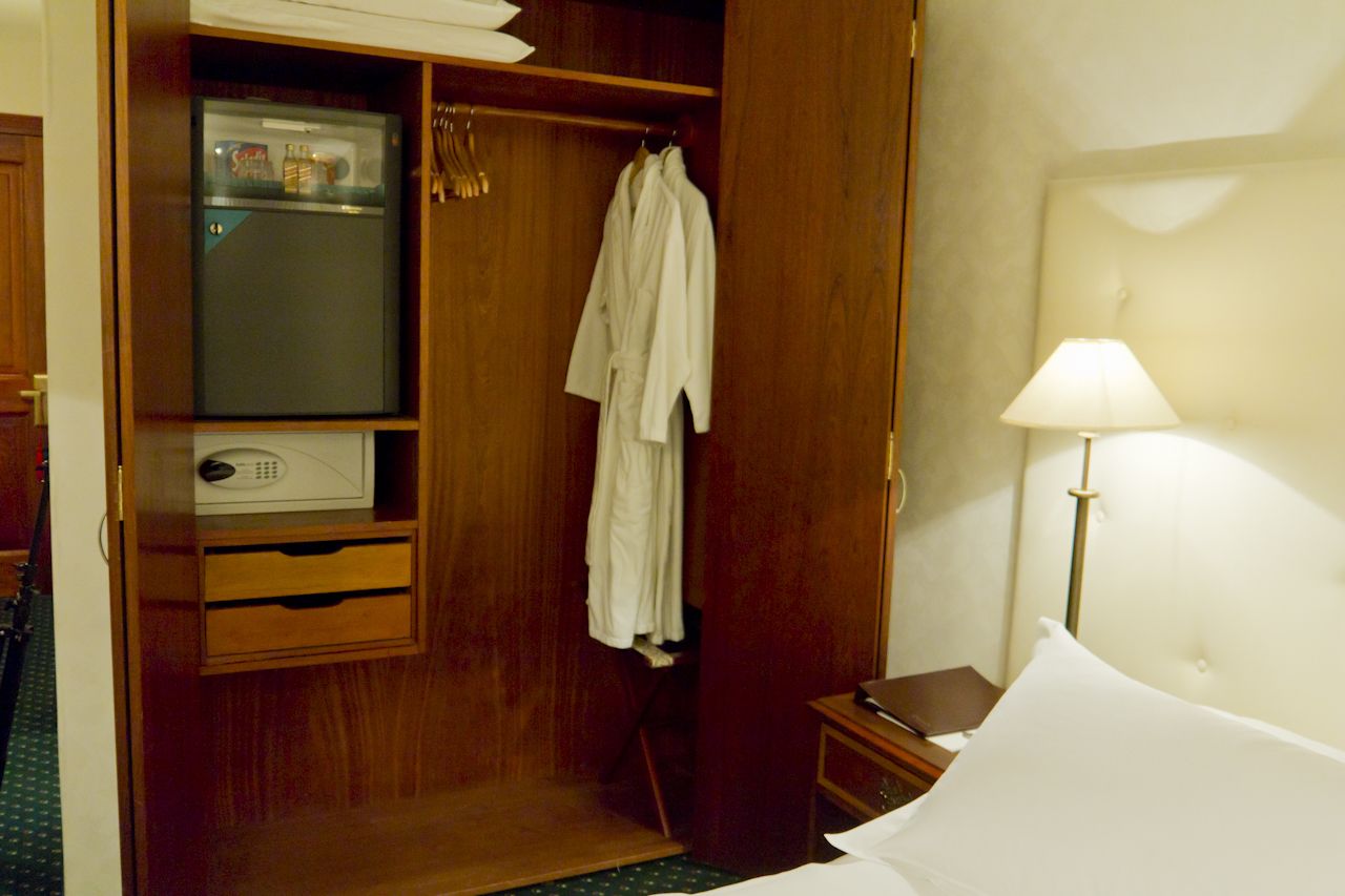 A hotel room with a wooden wardrobe, a small desk with a lamp, and a bed with white bedding.