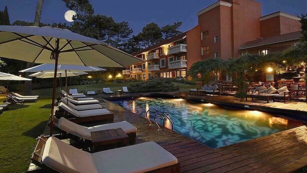 Zona de piscina con sillones de descanso y sombrillas, rodeada por un edificio con varios balcones.