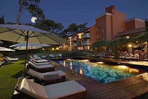 Zona de piscina con sillones de descanso y sombrillas, rodeada por un edificio con varios balcones.
