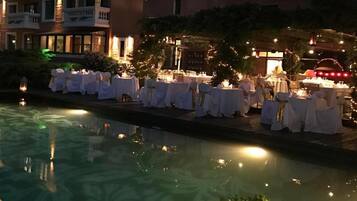 A poolside area with tables set for an event, surrounded by buildings with balconies and warm lighting.