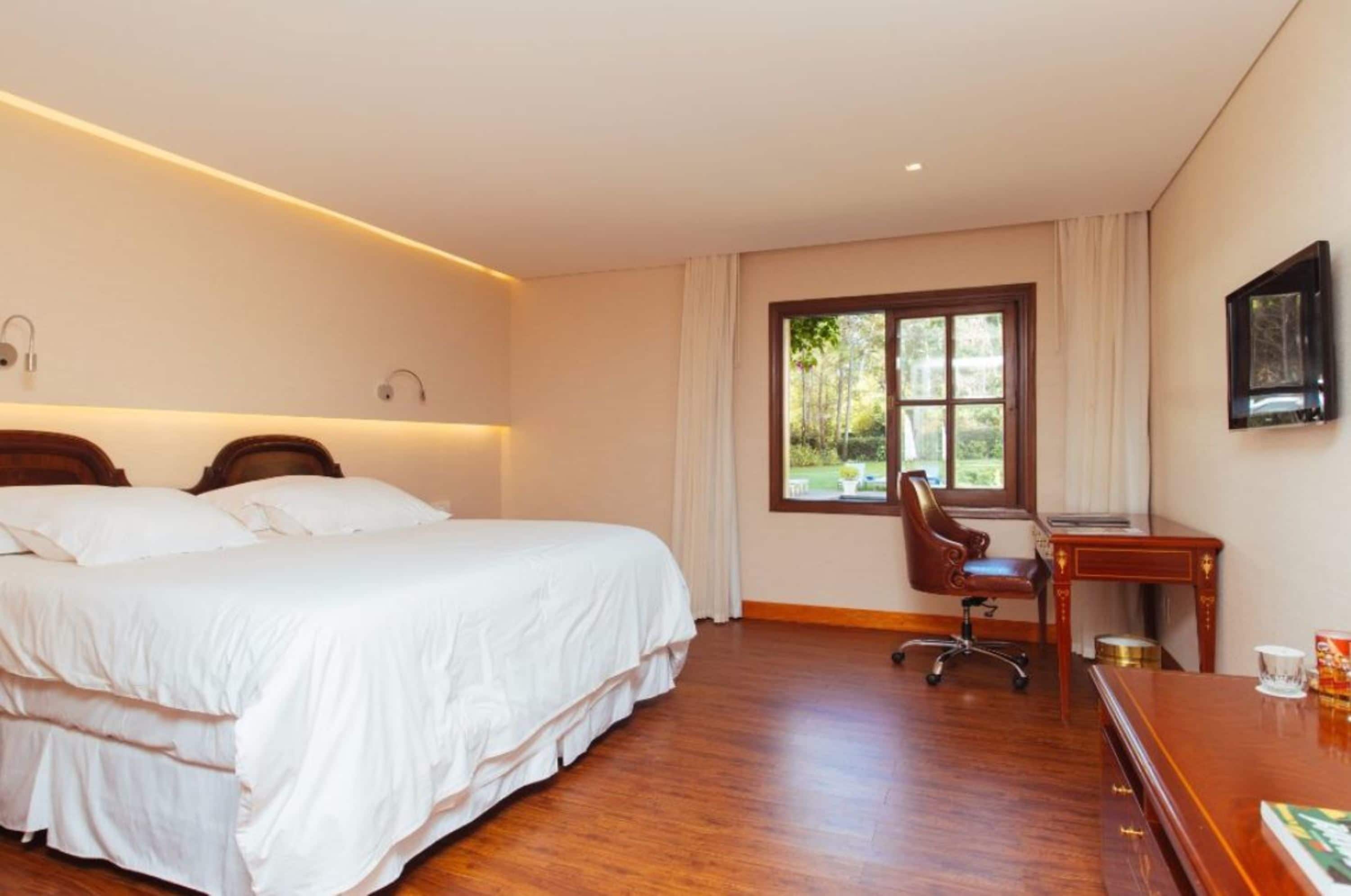 Habitación de hotel con una cama grande, un escritorio y una silla. Se ve un paisaje verde a través de la ventana.