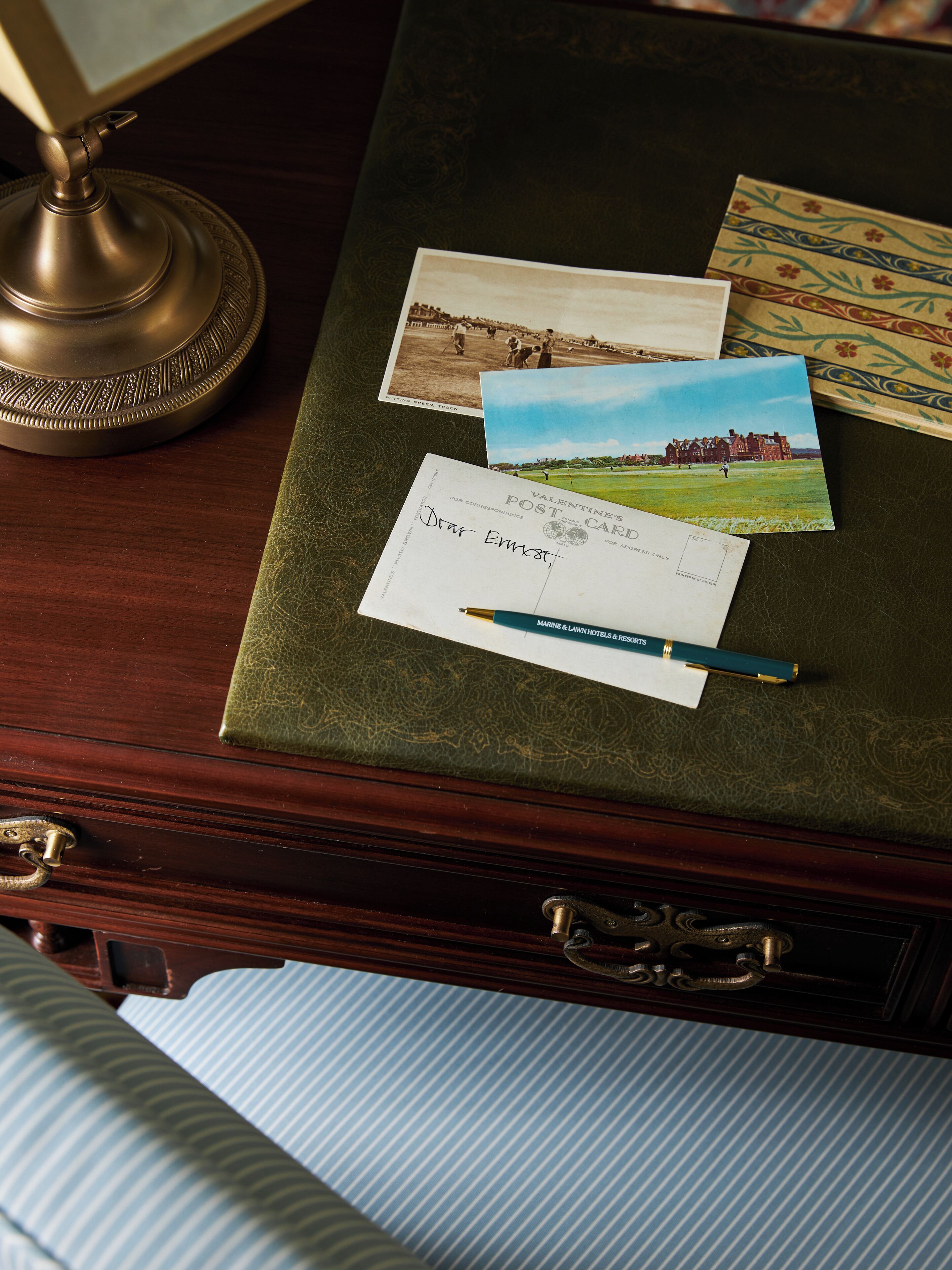 A desk with a brass lamp, a green book, a postcard, a handwritten note, and a pen.