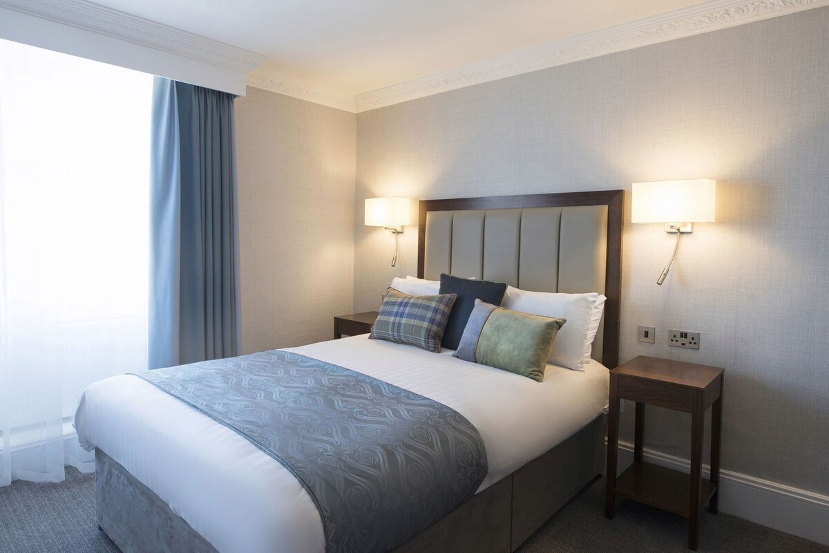A hotel room with a large bed, two bedside tables, a headboard, and two wall-mounted lamps.