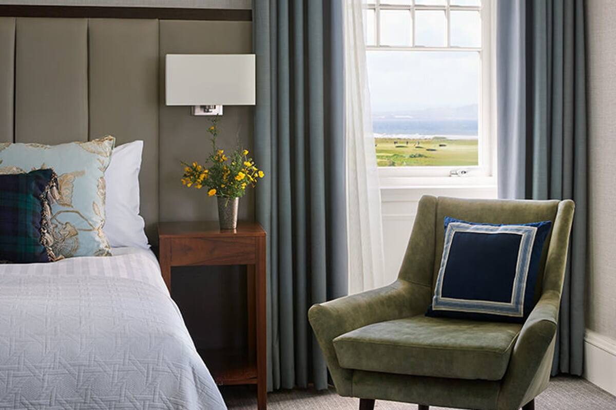 A hotel room with a bed, a chair, a nightstand with a lamp, a vase with flowers, and a window with curtains.